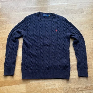 Mörkblå kabelstickad tröja Polo Ralph Lauren - Snygg mörkblå kabelstickad tröja från Polo Ralph Lauren med klassisk röd broderad logga på bröstet. Tröjan är inprincip oanvänd. Har du några ytterligare frågor är det bara ställa dom.