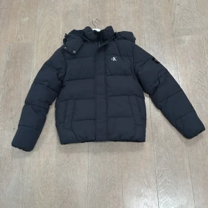 Svart pufferjacka Calvin Klein Jeans XS - Svart pufferjacka från Calvin Klein Jeans med broderad CK-logga på bröstet och patch på ärmen. Jackan har huva med tryckknappar, dragkedja framtill och quiltad design. det är storlek xs men den sitter som en vanlig s
