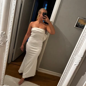 Klänning med rosett - Säljer en vit axelbandslös långklänning från Dress Decode i storlek M. Klänningen har en snygg rosett i ryggen och är gjord i glansigt satinmaterial. Den är figurnära och har dragkedja i sidan. Perfekt för fest, möhippa eller bröllop.