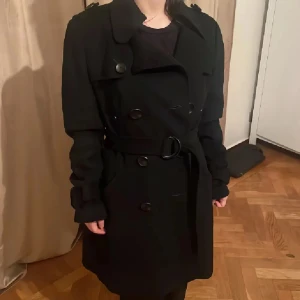 Svart ull trenchcoat med bälte och knappar - Snygg svart ullblend trenchcoat med klassisk dubbelknäppning, bred krage och bälte i midjan. Jackan har långa ärmar med dekorativa remmar och stora svarta knappar både fram och bak. Perfekt för dig som gillar stilrena och tidlösa plagg. Strl 38 men passar så fint på xs/s se bilderna😊 Perfekt till detta kalla väder 