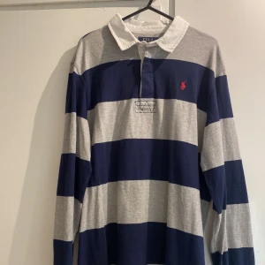 Randig pikétröja från Polo Ralph Lauren - Klassisk långärmad pikétröja från Polo Ralph Lauren i marinblått och grått med breda ränder. Tröjan har vit krage, knäppning vid halsen och röd broderad logga på bröstet. Perfekt för en sportig och avslappnad stil. Helt oanvänd.