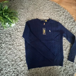 Marinblå stickad tröja Polo Ralph Lauren - Snygg marinblå stickad tröja från Polo Ralph Lauren med klassiskt kabelstickat mönster och broderad logga på bröstet. Rund halsringning och långa ärmar, perfekt för dig som gillar stilrena och tidlösa plagg.