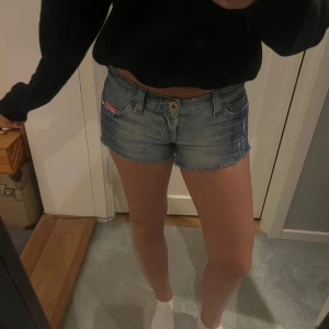 Lågmidjade jeansshorts med rosa detalj - Säljer ett par lågmidjade jeansshorts i klassisk blå denim med rå kant och slitningar. Shortsen har fem fickor, bälteshällor och en unik rosa detalj med tryck på bakfickan samt rosa sömmar. Perfekt för dig som gillar street och chill vibe.