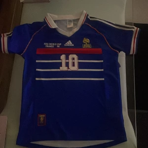Blå Frankrike Zidane VM 98 fotbollströja - Säljer en ikonisk blå Frankrike fotbollströja från VM 98 med nummer 10 och Zidane på ryggen. Adidas-logga, vita och röda detaljer, klassisk krage och korta ärmar. Flera vita ränder på framsidan och broderat landslagsmärke. Perfekt för samlare eller fans!