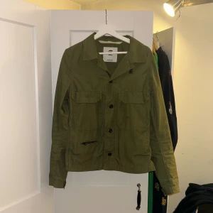 Grön overshirt från G-Star RAW - Snygg mörkgrön overshirt från G-Star RAW med klassisk krage, två stora bröstfickor med lock, knappstängning och en extra ficka med dragkedja. Perfekt att slänga över en hoodie eller t-shirt för en chill look. Är öppen för att gå ner i pris