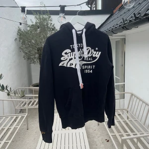 Svart Superdry hoodie med vit logga - Svart hoodie från Superdry med stor vit logga och text på bröstet. Tröjan har huva med vita snören, känguruficka och ribbade muddar. Perfekt för chill dagar och har en klassisk streetstil. Materialet är mjuk bomull och polyesterblandning.