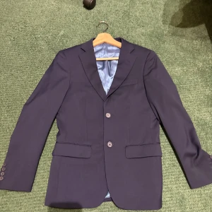 Mörkblå kavaj med knappar - Snygg mörkblå kavaj med klassisk passform och två knappar framtill. Kavajen har fickor med lock och slät yta, samt ljusblått foder på insidan. Perfekt för dig som vill ha en stilren look.