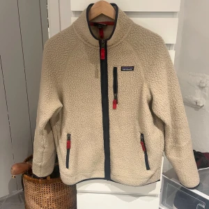 Patagonia fleece Biege - Säljer en beige fleecejacka från Patagonia med mörkblå detaljer och röda dragkedjor. Jackan har hög krage, tre fickor med dragkedja och en mysig teddystruktur. Perfekt för kalla dagar och chill stunder. Snygg och enkel att matcha med jeans eller mjukis.