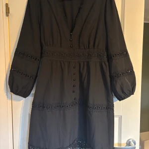 Svart klänning med spetsdetaljer - Svart klänning med v-ringning, dekorativa knappar framtill och breda spetsdetaljer vid midja, ärmar och nederkant. Klänningen har långärmade ballongärmar med resår och är tillverkad i ett mjukt bomullstyg. Perfekt för dig som gillar romantiska detaljer och en feminin look.