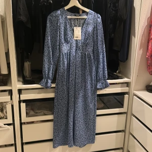 Blommig blå långklänning från Monki - Supersöt långklänning från Monki i ljusblått med svart blommigt mönster. Klänningen har v-ringning, långa ärmar med volangdetalj vid ärmslut och en avslappnad passform. Perfekt för dig som gillar romantiska vibes och vill sticka ut lite extra.