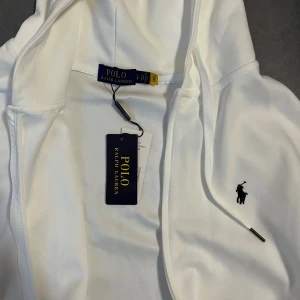 Vit hoodie från Polo Ralph Lauren - Vit hoodie från Polo Ralph Lauren med klassisk svart logobrodyr på bröstet. Tröjan har huva med snörning, dragkedja framtill och två fickor. Mjuk bomullskvalitet och stilren design som passar perfekt till jeans eller joggers.