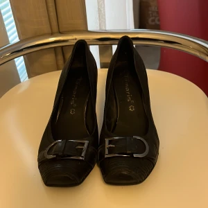 Svarta pumps från Tamaris med spänne - Snygga svarta pumps från Tamaris med rundad klack och markerad fyrkantig tå. Skorna har ett dekorativt metallspänne framtill och är tillverkade i skinn med diskret textur. Perfekt för dig som gillar klassisk stil med en twist.