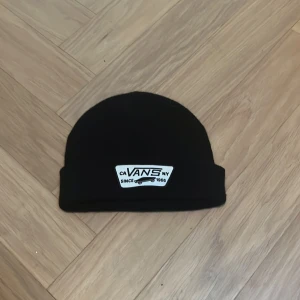 Svart Vans mössa - Svart mössa från Vans med klassisk uppvikt kant och vit patch framtill med logga och skateboardmotiv. 