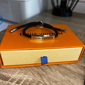 Louis Vuitton svart armband i silver - Stilrent svart armband från Louis Vuitton med justerbar textilrem och silverfärgade metalldetaljer. Armbandet har en platta med Louis Vuitton-loggan ingraverad samt en rund detalj med LV-emblemet. Perfekt accessoar för en trendig look.