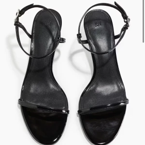Svarta sandaler med klack från H&M - ALDRIG ANVÄNDA. Snygga svarta sandaler från H&M med smala remmar och justerbart spänne runt ankeln. Skorna har låg klack och en stilren, minimalistisk design med glansig finish. Perfekta för dig som gillar enkel men trendig stil. Rund tå och öppen tå för en fräsch look.