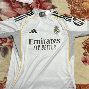 Real Madrid Mbappé 10 fotbollströja - Säljer en officiell Real Madrid fotbollströja från Adidas med Mbappé och nummer 10 på ryggen. Tröjan är vit med svarta och gula detaljer, korta ärmar och tillverkad i lätt polyester med Aeroready-teknologi. Klubbmärke och sponsortryck på bröstet.