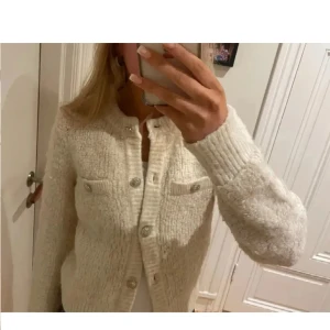 Beige stickad kofta med knappar - Supermysig beige stickad kofta med glittriga knappar och två fejkade bröstfickor. Koftan har rund halsringning, ribbade muddar och lång ärm. Perfekt att slänga över en topp för en chill och snygg look.