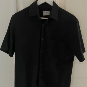 Svart kortärmad skjorta NN07 - Stilren svart skjorta från NN07 med klassisk krage, korta ärmar och bröstficka. Skjortan har knappar hela vägen och är perfekt för dig som gillar enkel och clean stil. Materialet känns mjukt och lätt mot huden. Nypris 1400, S passar M
