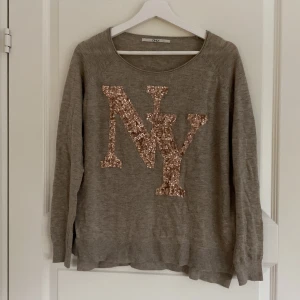 Beige tröja med paljett-NY från ONLY - Beige långärmad tröja från ONLY med stor NY-logga i glittriga paljetter framtill. Tröjan har rund halsringning och är i mjukt material, perfekt för en chill och snygg look. Passar dig som gillar lite extra bling i vardagen.
