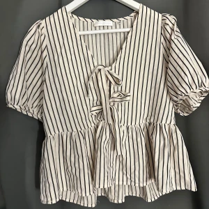 Randig blus från Carin Wester, stl 34 - Supersöt blus från Carin Wester i vitt med svarta vertikala ränder. Modellen har v-ringning, knytband framtill, puffiga korta ärmar och volang nedtill. Perfekt för dig som gillar en luftig och trendig look med extra detaljer💓