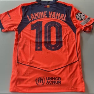 FC Barcelona Lamine Yamal tröja Nike - Säljer en FC Barcelona fotbollströja från Nike med Lamine Yamal och nummer 10 på ryggen. Tröjan är orange med mörkblå detaljer, kortärmad och har Champions League och UEFA Foundation patches på ärmen. Materialet är lätt och andas, perfekt för match eller träning. Replika, endast handtvätt gäller!
