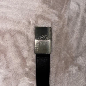 Svart bälte från Tiger of Sweden  - Snyggt svart bälte i skinn med ett rektangulärt silverfärgat spänne. Spännet har en graverad ros som ger en unik och cool detalj. Perfekt accessoar för att lyfta din outfit och ge den lite edge. Markaden nu är 1000-1200kr/ säljer den här för 650kr men pris kan diskuteras 