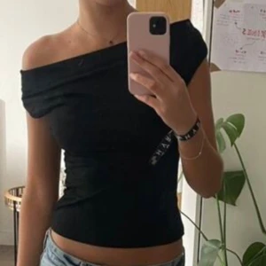 Svart offshoulder topp med knyt - Säljer en svart offshoulder topp i stretchigt material med snygg knytning i ryggen. Toppen är figurnära och har en cool asymmetrisk axel, perfekt för att sticka ut. Går att justera och ha påde med djup ringning framtill eller off shoulder beroende på hur man knyter den baktill. Normal i storlek. 