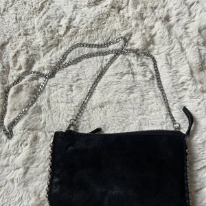 Svart axelväska med kedja och pärldetaljer - Snygg svart axelväska i mocka med silverfärgad kedjerem och dragkedja. 
