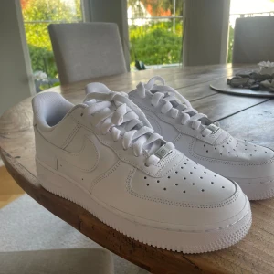 Nike Air Force 1 helvita sneakers - Klassiska Nike Air Force 1 sneakers i helvitt skinn med vit sula och snörning. Modellen har perforerade detaljer på tån för extra ventilation och en chunky, platt sula. Perfekta för dig som gillar en clean och tidlös look.helt nya aldrig använda 
