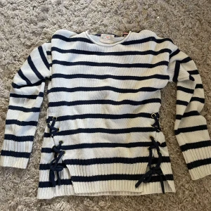 Randig stickad tröja med snörning - Säljer en stickad tröja från Holly & Whyte i vitt och mörkblått med breda ränder. Tröjan har en cool snörningsdetalj på sidorna. Den är väldigt stor i storleken då den är 134/140 men passar mig som vanligtvis har S i tröjor