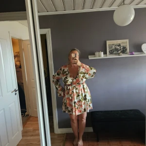 Blommig omlottklänning från Boohoo - Säljer en vit omlottklänning från Boohoo med blommigt mönster i rosa och grönt. Klänningen har långärmade ärmar, v-ringad hals och ett knytband i midjan för extra snygg passform. Perfekt för dig som gillar romantiska vibes och vill sticka ut.