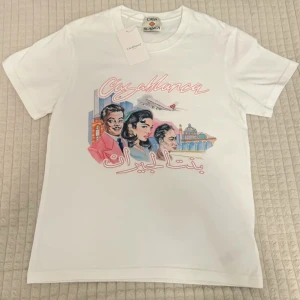 Vit Casablanca t-shirt med tryck - Vit t-shirt från Casablanca med färgglatt tryck framtill som visar personer, byggnader och ett flygplan. T-shirten har rund hals och korta ärmar. Materialet är mjuk bomull och trycket har en retrovibe med rosa och blå toner samt text på arabiska.