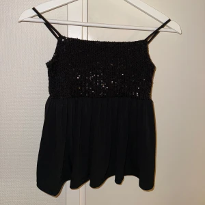 Svart paljettlinne med tunna axelband - Säljer ett svart linne med tunna axelband och glittriga paljetter upptill. Toppen har en smockad överdel med paljetter och ett flowy, lätt tyg nedtill. Perfekt för dig som vill sticka ut med lite extra bling. Snygg till både jeans och kjol!