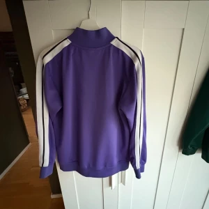 Lila Palm Angels track jacket - Säljer en lila track jacket från Palm Angels med vita ränder längs ärmarna och vit dragkedja. Tröjan har hög krage, två fickor med dragkedja och logga på bröstet. Perfekt för en sportig och trendig look.