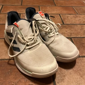 Vita Adidas sneakers med svarta ränder - Säljer ett par fräscha vita Adidas sneakers med klassiska svarta ränder på sidorna och orange detalj på plösen. Skorna har snörning, platt sula och är tillverkade i syntetmaterial med mesh och skinnliknande detaljer. Perfekta för träning eller sportiga outfits.