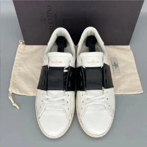 Valentino Garavani Open sneakers vit/svart - Snygga Valentino Garavani Open sneakers i vitt skinn med bred svart läderdetalj över snörningen. Klassisk rund tå, vita snören och ikoniska nitar på hälen. Perfekta för dig som gillar stilrena och lyxiga sneakers med en twist.