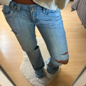 Ljusblå baggy jeans med slitningar - Säljer ett par ljusblå jeans med baggy passform och slitna detaljer vid knäna. Jeansen har klassisk femficksdesign, dubbla knappar i midjan och coola sömmar vid fickorna. Materialet är jeans och de har en avslappnad vibe. De är slitna längst ner men annars inga andra defekter 