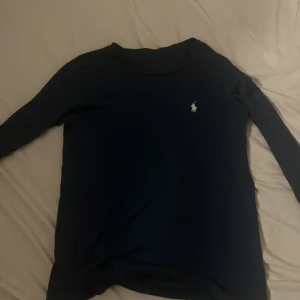 Mörkblå långärmad tröja från Ralph Lauren - Snygg mörkblå långärmad tröja från Ralph Lauren med klassisk vit broderad logga på bröstet. Tröjan har en enkel och stilren design med rund halsringning och är tillverkad i mjukt bomullsmaterial. Perfekt för en clean och tidlös look. Vet dock ej storlek men jag som 155 är den lite stor och sitter inte tajt men sitter bra vid ärmen. Pris kommer kunna sänkas