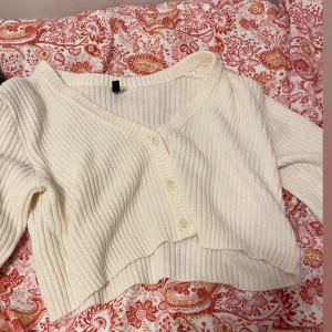 Vit ribbad croppad bolerotopp - Superfin vit ribbad bolerotopp med v-ringning och knappar framtill. Croppad modell som ger en trendig look och passar perfekt till höga jeans eller kjol. Mjuk och stickad känsla som är både bekväm och snygg.