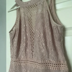 Beige spetsklänning med öppen rygg - Supersnygg beige spetsklänning med blommigt mönster och detaljerad spets vid midjan. Klänningen är ärmlös, har en öppen rygg och ett coolt rutigt meshparti framtill. Passformen är slim och längden är kort, perfekt för dig som gillar en chic och feminin stil.