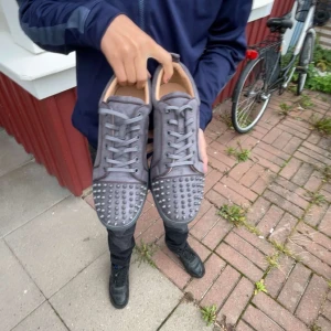 Grå mocka sneakers från Louboutin - Säljer ett par grå sneakers från Louboutin i mocka med coola nitar framtill och klassisk röd sula. Skorna har snörning och rund tå, samt en platt sula. Perfekta för dig som vill sticka ut med en lyxig och edgy stil.
