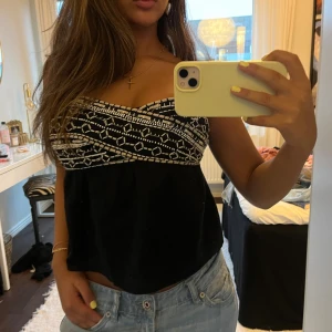 Svart och vit tubtopp med mönster - Säljer en svart tubtopp med vit broderad detalj upptill. Toppen är axelbandslös och har en lätt utsvängd passform nedtill. Mönstret upptill är små stenar, broderi och paljetter. Liten för att va en M