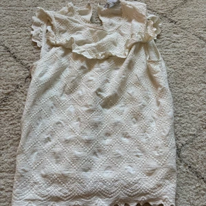 Blus från Other Stories - Supersöt vit kortklänning från & Other Stories med broderade detaljer och volang vid axlarna. Klänningen har ett romantiskt mönster och är ärmlös, perfekt för varma dagar. Tillverkad i bomull för en luftig känsla.