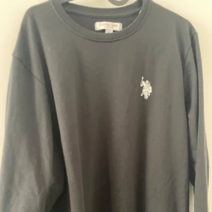 Svart sweatshirt från U.S. Polo Assn. - Svart långärmad sweatshirt från U.S. Polo Assn. med klassisk rund hals och broderad logga på bröstet. Tröjan är i bomull och har en enkel, stilren design som passar perfekt till jeans eller joggers.