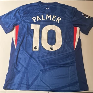 Chelsea Palmer 10 Nike matchtröja blå - Chelsea FC hemmatröja med Palmer och nummer 10 på ryggen. Tröjan är blå med vita detaljer, har klubbmärke och Nike-logga på bröstet samt Premier League-märke på ärmen. Tillverkad i lätt och ventilerande material, perfekt för fotboll eller supporterliv. Replika, endast handtvätt gäller!