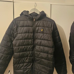 Svart pufferjacka Lyle & Scott Junior - Svart pufferjacka från Lyle & Scott Junior med huva och gul logga på bröstet. Jackan har horisontella sömmar, dragkedja framtill och innerficka. Perfekt för kyliga dagar och riktigt snygg streetstil.
