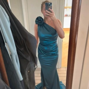 Blå långklänning med oneshoulder och rosett - Säljer en glansig blå långklänning i satin med oneshoulder och stor rosett på axeln. Klänningen har figurnära passform, draperade detaljer och slits på sidan. Perfekt för bal eller andra tillställningar. Vet tyvärr inte vad de är för märke på den då jag köpte den second hand. Har lite defekter i tyget men inget som påverkar helhets intyget. 