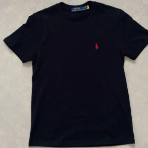 Svart t-shirt från Polo Ralph Lauren -  Mer som S. Svart t-shirt från Polo Ralph Lauren med klassisk röd broderad logga på bröstet. Modellen är kortärmad och har rund hals. Tillverkad i mjuk bomull som känns skön mot huden. Perfekt för dig som gillar stilrena och tidlösa plagg.