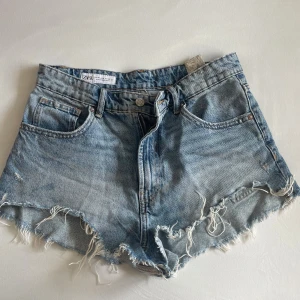 Blå jeansshorts med fransig kant Zara - Snygga blå jeansshorts från Zara med hög midja och rå, fransig kant nertill. Klassisk femficksmodell med dragkedja och knapp framtill. Perfekta för sommaren och ger en avslappnad vibe till din outfit.