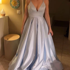 klänning - Säljer en ljusblå långklänning med djup v-ringning och tunna axelband. Klänningen har en voluminös kjol med mycket tyg som ger en elegant siluett. Perfekt för bal eller student. Materialet är glansigt och känns lyxigt.